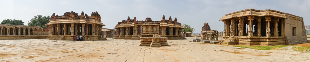 Fototapeta premium Hampi, Karnataka, India - November 5, 2013: Vijaya Vitthala Temple. Panorama shot of main brown stone buildings under light blue sky and beige stone courtyard uip front. People add color.