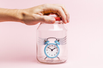 person holding alarm clock with origami wings at closed jar. pink pastel background. catch time concept. hope conceptual