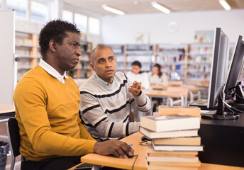 Portrait of two adult men engaged in research, sitting at table with computer and books, finding information in library