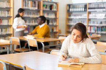 Interested young Latina spending time in library, reading books and making notes. Self-education concept