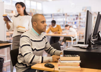 Young adult man sitting at table with computer and books, finding information at library