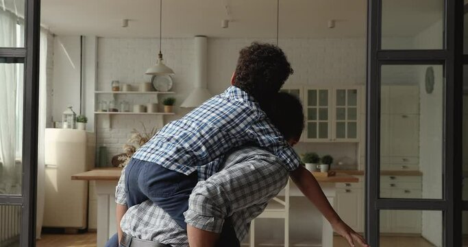 African kid boy piggyback ride on daddy back play together feel happy. Caring American father run in living room holding little 5s son on his back. Weekend leisure games with children at home concept
