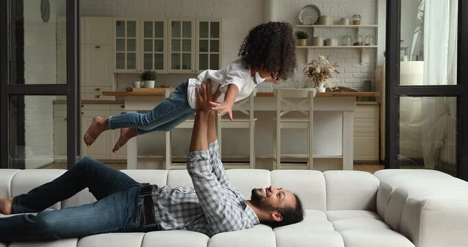 Happy young african mixed race dad lying on sofa lifting in outstretched arms preschool daughter, joyful kid little 4s girl imagines like she is airplane play active games with loving father at home