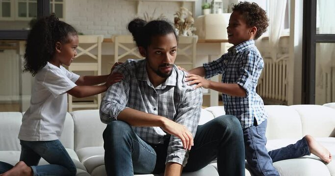 Hyperactive african 5s son and daughter want to play with tired father sit on sofa feels exhausted to spend time with children. Upbringing difficulties, need repose, divorced helpless parent concept