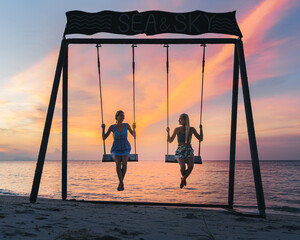 Two happy smiling young caucasian woman with blond long hair swinging on a swing standing by the...