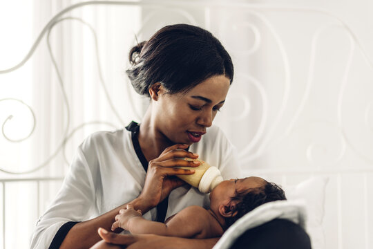 Portrait Of Enjoy Happy Love Family African American Mother Playing With Adorable Little African American Baby.Mom Feeding Bottle Of Milk To Baby Cute Son In A White  Bedroom.Love Of Black Family 