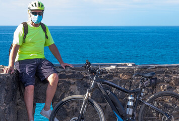 Coronavirus. Portrait of a senior white haired man with bicycle and helmet doing sport activity enjoying nature and sea, wearing a protective face mask due to coronavirus contagion