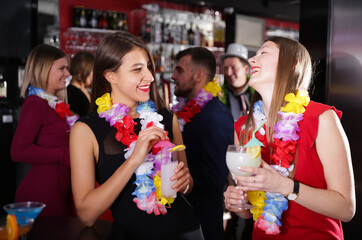 Cheerful girls dancing and toasting drinks on Hawaiian party in bar