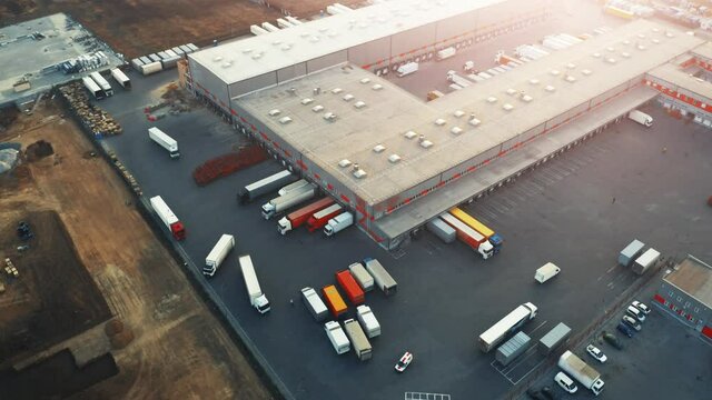 Logistics park with warehouses, loading hub and a lot of semi-trailer trucks await for load and unload goods from cargo containers at ramps. Aerial view at sunset