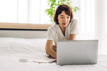 Smiling happy asian woman relax using laptop computer working and talk with group of multiracial business diverse colleagues discussing meeting strategy sharing ideas on bed at home.work from home 