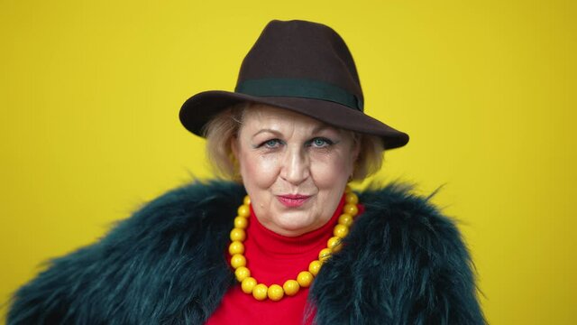 Close-up of confident flirty senior woman in elegant hat looking at camera smiling. Portrait of coquette Caucasian lady posing on yellow background. Flirt and beauty concept.