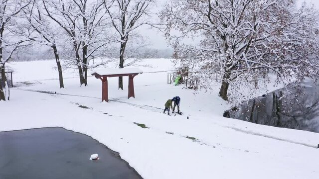 Men cleaners holding special long shovels clean paths in park covered with heavy snow near lakes among bare trees