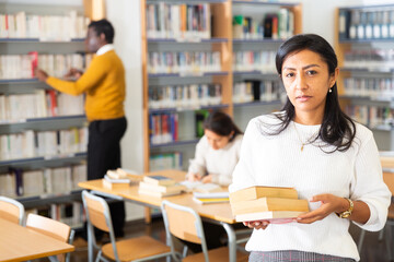 Confident latin american adult woman returning pile of books to librarian in public library