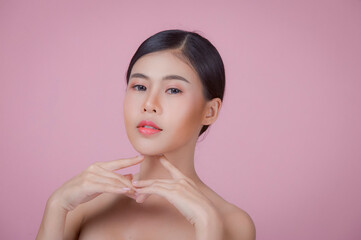 Youthful smiling Asian woman with hand touching face isolated on pink background for beauty and skin care concepts