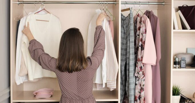 Female trying to picks up an outfit in home wardrobe. Young woman is choosing clothes on a racks in closet searching what to wear, back view. Concept of everyday dilemma in outfit choosing.
