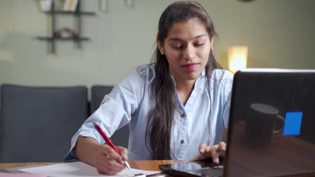 Young business woman writing down notes by looking laptop - concept of employee or student online training class at home