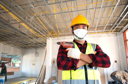 A young Asian construction worker in safety clothing and work gloves is fastening the drywall ceiling to the metal frame using an electric screwdriver on the ceiling covered in shiny aluminum foil.