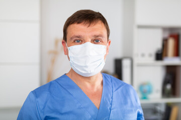 Fototapeta na wymiar Portrait of confident adult male doctor in blue uniform and disposable face mask in modern medical office..
