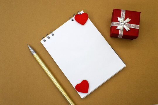 A notebook with a blank white sheet and two red hearts lies between a red gift box and a yellow pen. Concept photo