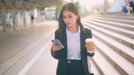 A young business woman wearing black suit is using smart phone , holding a cup of coffee in the...