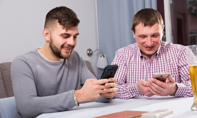 Positive men sitting at home table absorbedly looking at phones