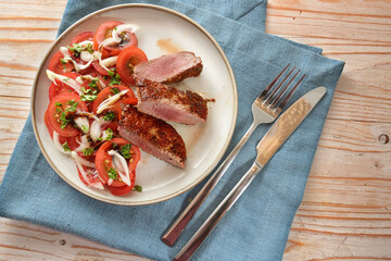 Roasted lamb meat, tomato salad, onions and parsley garnish on a plate and a blue napkin with cutlery on a rustic wooden table, high angle view from above, selected focus
