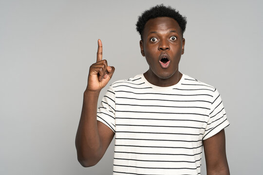 Stupefied afro young man makes big eyes opens mouth widely, raise finger up, surprised by suddenly invented a great idea and looking at camera, wear stripped t-shirt isolated on studio grey background