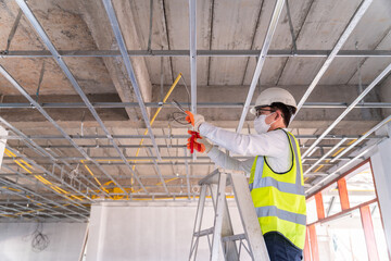 Asian handsome electrician wearing a mask installing laying electrical cables on the ceiling with pliers inside the house under construction.