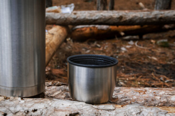 Naklejka na ściany i meble Thermos of tea in the forest close-up. Metal thermos with a mug in selective focus. One-day hike in winter in the mountains and forest.A place to relax.The concept of hiking, tourism, active lifestyle