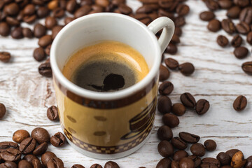 Cup of espresso surrounded with a lot of roasted coffee beans on a white wooden background.