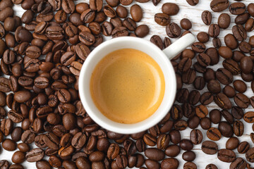 Cup of espresso surrounded with a lot of roasted coffee beans on a white wooden background.