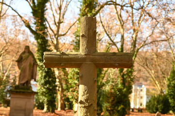A stone cross at a graveyard during autumn. Brown leaves are lieing on the soil. A statue of the Virgin Mary is in the background.