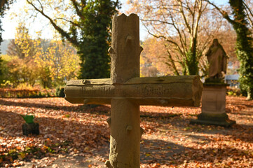 A stone cross at a graveyard during autumn. Brown leaves are lieing on the soil. A statue of the Virgin Mary is in the background.