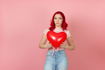 a resentful, upset young woman with red hair holds a large flying red heart-shaped balloon in her hands , isolated on a pink background.