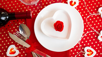 A white plate with a knife and fork on a bright red background with wine bottle. Heart shaped white plate. Valentines Day. The view from the top.