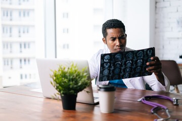 Side view of African American black male doctor in white coat analyzing history disease of patient using MRI brain head scan, working on laptop. Concept of medicine and health care.