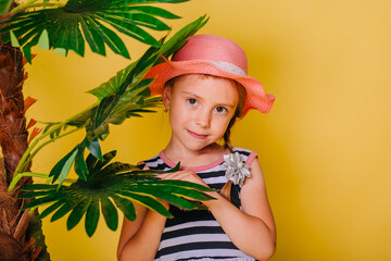 A little girl in a pink hat and a striped dress stands next to a palm tree on a yellow background.