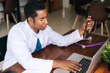 Top view of African American black male doctor in white coat analyzing history disease of patient using MRI brain head scan, working on laptop. Concept of medicine and health care.