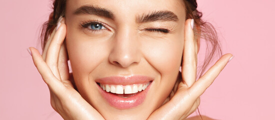 Skincare and beauty. Close up of smiling young woman winking at camera, touching shiny healthy skin, nourished face after tea tree and lemon facial serum and moisturizer, pink background - obrazy, fototapety, plakaty