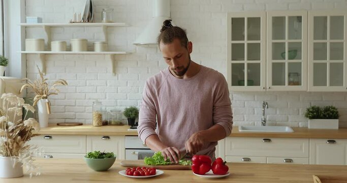 Young single vegan man with tied hair standing alone in domestic kitchen holding knife cutting fresh lettuce for vegetable salad, prepare dinner for romantic date. Healthy eating, vegetarian lifestyle