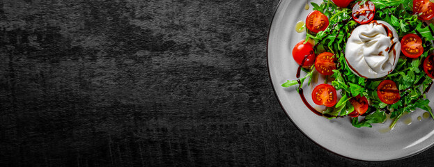 salad with burrata cheese, arugula salad and tomatoes on plate on Dark grey black slate background