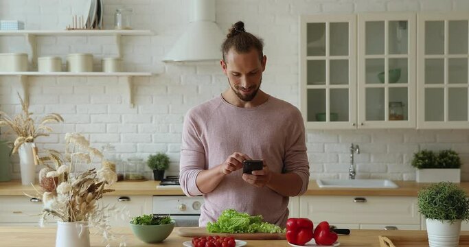Young single man holding smartphone search healthy food recipes on-line, use easy meal ideas application, distracted from vegetable salad preparation chatting online. Concept of cookery, mobile app