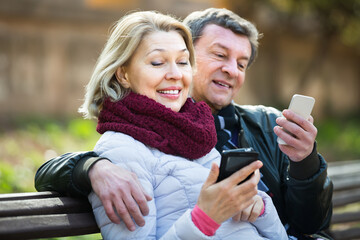 Joyful smiling elderly couple exchanging phone numbers during outdoor date.