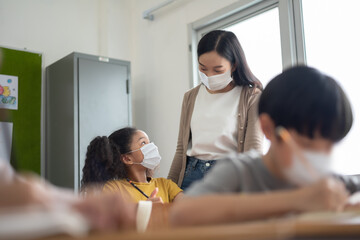Young Asian Teacher with African-American girl wearing protective face masks studying in classroom