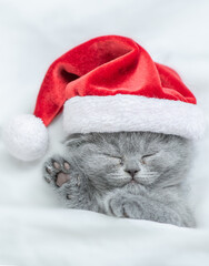 Funny kitten wearing red santa's hat sleeps under a white blanket on a bed. Top down view