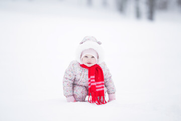 One smiling baby girl in pink overalls and red scarf squatting on white deep snow and enjoying beautiful winter day at park. Front view.