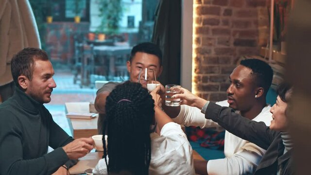 Diverse happy friends clinking glasses and cups with different beverages while celebrating something in restaurant. Friends toasting while enjoying dinner together in trendy cafe or restaurant