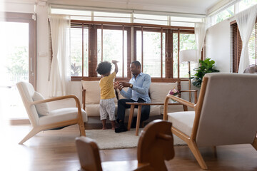 African American Dad playing and having fun with his little son on the couch of living room at home.