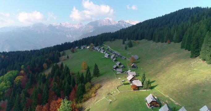 Aerial view of small Alpine village in Slovenia. Drone flying over spruce forest and mountain cabins. Alps mountains in the distance. Beautiful and colorful trees in autumn. Left truck, parallel