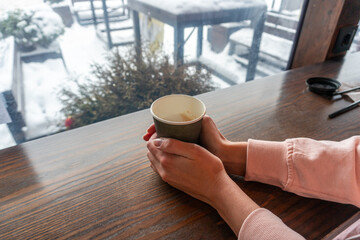 A girl with two hands holds a paper cup with coffee in a cafe overlooking a winter street